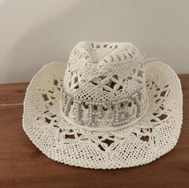 Puede incluir: Un sombrero de vaquero blanco de ganchillo con una banda decorativa que deletrea "WIFEY" en letras plateadas y perlas de imitaci&oacute;n. El sombrero tiene un ala ancha y un patr&oacute;n floral.