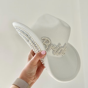 May include: White cowboy hat decorated with pearls and rhinestones. The word "Bride" is spelled out in pearls. The brim is lined with pearls and rhinestones. A fashionable accessory for a wedding or hen party.