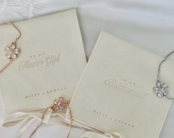 Könnte beinhalten: Zwei cremefarbene Beutel mit den Aufschriften "TO MY Flower Girl" und "TO MY Bridesmaid". Blumenförmige Armbänder sind zu sehen. Die Beutel stammen von "Gifty London".