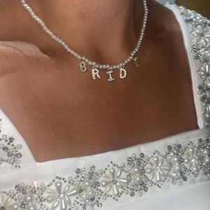 May include: A pearl necklace with gold-colored letters spelling "BRIDE". The necklace is worn over a white dress with pearl and crystal embellishments. The necklace is a bridal accessory.