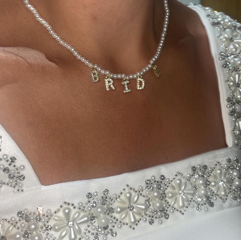 May include: A pearl necklace with gold-colored letters spelling "BRIDE". The necklace is worn over a white dress with pearl and crystal embellishments. The necklace is a bridal accessory.