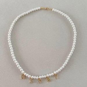 May include: A white pearl necklace with gold-colored clasps and charms spelling out the word "WIFEY". The necklace is made of small, round, white pearls strung together. The charms are encrusted with small, clear crystals.
