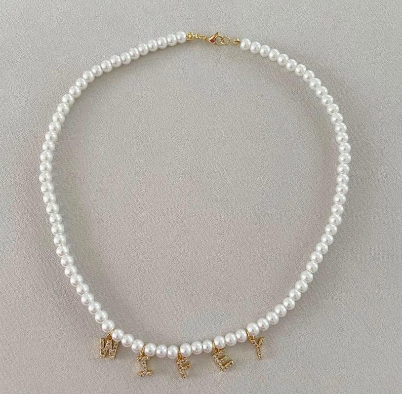 May include: A white pearl necklace with gold-colored clasps and charms spelling out the word "WIFEY". The necklace is made of small, round, white pearls strung together. The charms are encrusted with small, clear crystals.