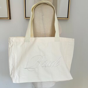 May include: Cream-colored tote bag with the word "Bride" embroidered in a delicate script. The bag has sturdy handles and is made of a soft, durable fabric. Perfect for carrying essentials on the wedding day or as a bridal shower gift.