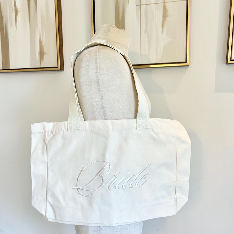 May include: Off-white canvas tote bag with the word "Bride" embroidered in a cursive script. The bag has two sturdy handles and is displayed on a mannequin. The background features framed artwork.