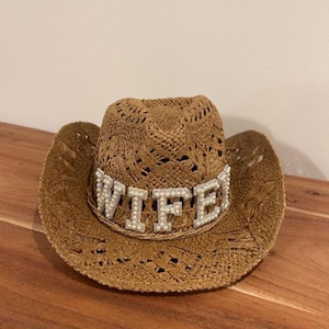 Puede incluir: Un sombrero de vaquero tejido marr&oacute;n con una banda decorativa que deletrea la palabra "WIFE" en letras plateadas con detalles de perlas. El sombrero tiene un ala ancha y un patr&oacute;n de tejido abierto decorativo.