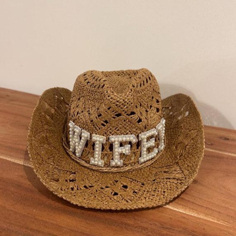 Puede incluir: Un sombrero de vaquero tejido marr&oacute;n con una banda decorativa que deletrea la palabra "WIFE" en letras plateadas con detalles de perlas. El sombrero tiene un ala ancha y un patr&oacute;n de tejido abierto decorativo.