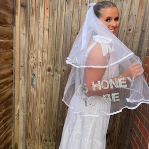 Puede incluir: Un velo de novia blanco con el texto "MRS HONEY TO BE" en letras plateadas con purpurina. El velo es de varias capas y tiene un detalle de lazo. La novia lleva un vestido blanco.