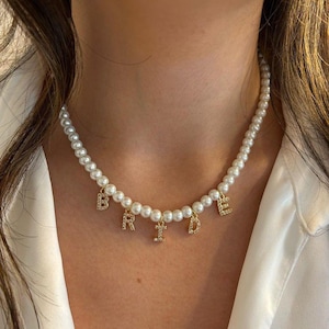 May include: A pearl necklace with gold-colored letters spelling "BRIDE". The letters are embellished with small, clear crystals. The necklace is composed of numerous small, round, white pearls.
