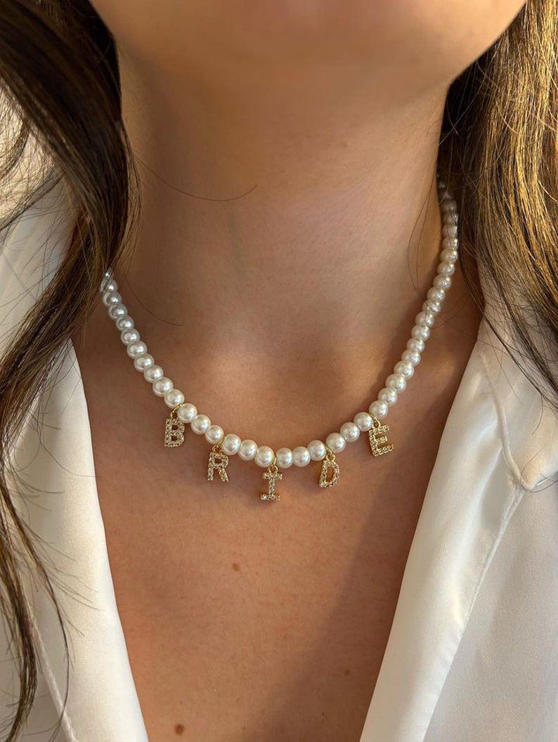 May include: A pearl necklace with gold-colored letters spelling "BRIDE". The letters are embellished with small, clear crystals. The necklace is composed of numerous small, round, white pearls.
