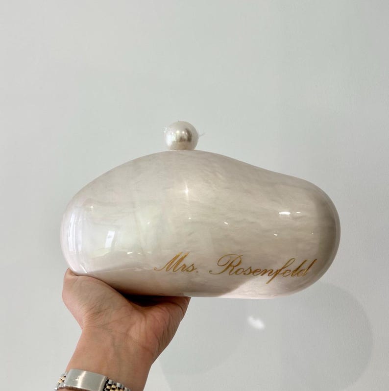 May include: A pearlescent, off-white clutch with a rounded shape and a small, pearl-like embellishment on top. The text "Mrs. Rosenfeld!" is inscribed in gold script on the front.