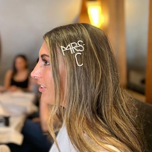 May include: A silver hair clip spelling out "MRS C" in rhinestones. The clip is set in the hair of a person with long, light brown hair. The background is blurred, suggesting an indoor setting, possibly a wedding or bridal event.
