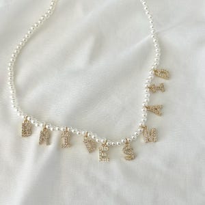 May include: A pearl necklace with gold-colored, rhinestone-studded letter charms spelling "BRIDESMAID". The necklace is on a white background. The letters are in a serif font and are evenly spaced along the necklace.
