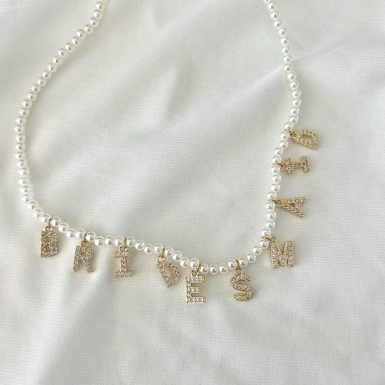 May include: A pearl necklace with gold-colored, rhinestone-studded letter charms spelling "BRIDESMAID". The necklace is on a white background. The letters are in a serif font and are evenly spaced along the necklace.