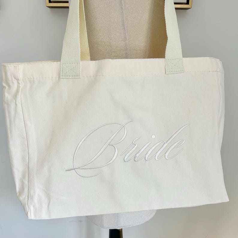 May include: Cream-colored tote bag with the word "Bride" embroidered in a delicate script. The bag has sturdy cream-colored handles and is made of a durable fabric, perfect for carrying essentials.
