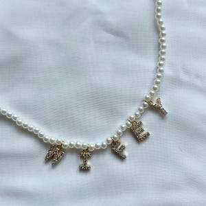 May include: A pearl necklace with gold-colored, rhinestone-studded letter charms spelling out the word "WILEY". The necklace features small, round pearls strung together, with the letters hanging below.