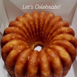 Può includere: Una torta a ciambella dorata con glassa lucida, presentata in una scatola bianca. La torta ha un design scanalato e un foro al centro. Il testo "Let's Celebrate!" è scritto in bianco sopra la torta.