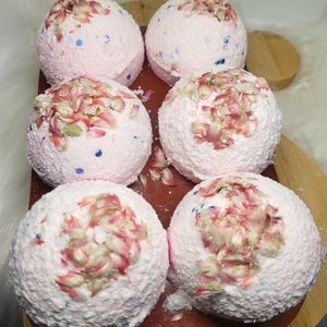May include: Six pink bath bombs with a textured surface and floral accents are arranged on a wooden board. The bath bombs have blue and pink speckles and are topped with dried flower petals. The board sits on a white surface.