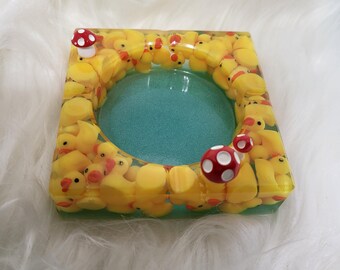 May include: Square resin ashtray filled with yellow rubber duckies and a turquoise center. Red and white mushroom accents are placed on the corners. The ashtray is on a white, fluffy surface.