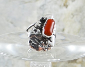 May include: A sterling silver ring featuring a textured, organic design. The ring has a prominent, rectangular, vibrant red coral stone set in a silver bezel. The ring's design is inspired by natural forms.