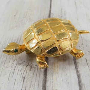 May include: A gold-toned turtle brooch with green gemstone eyes. The turtle's shell is textured with a grid pattern.