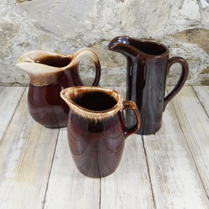May include: Three brown and cream glazed ceramic pitchers of varying sizes. The pitchers have a rustic, handmade look.