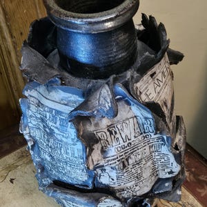 May include: A handcrafted ceramic vase with a dark, textured finish. The vase features a cylindrical body with newspaper-like designs and the text "REWAR" and "THE KID". The top is a dark, open cylinder.