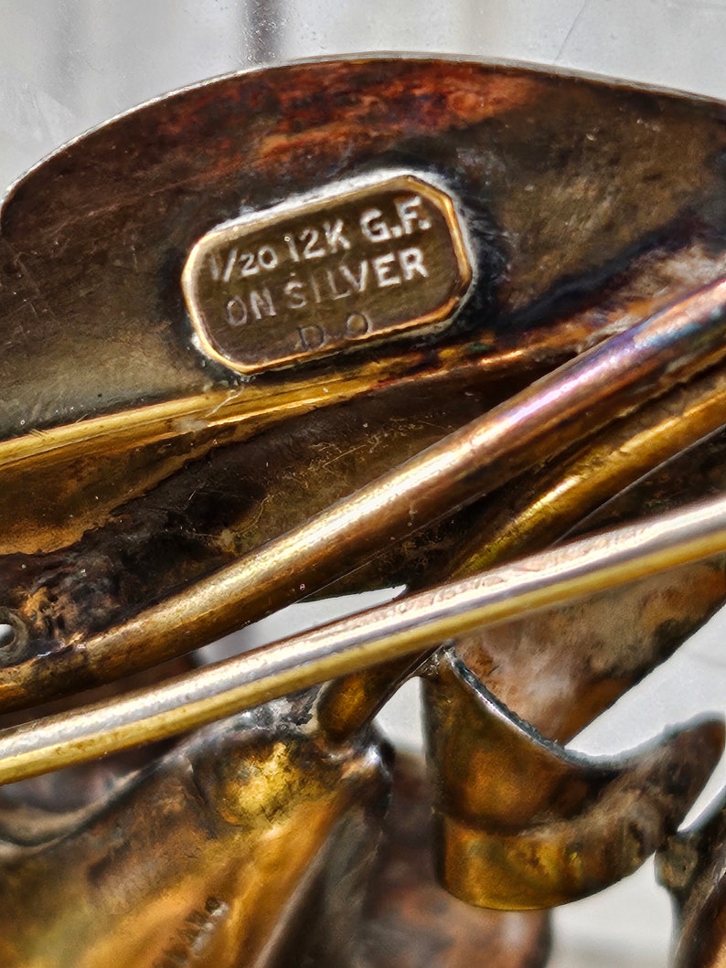 May include: Close-up of a gold-toned metal brooch with a detailed design. The brooch has a label that reads "1/20 12K G.F. ON SILVER D.O."
