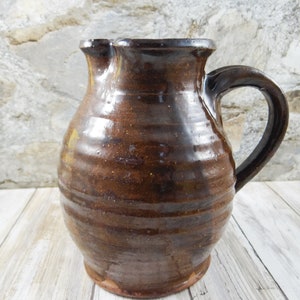 May include: A handmade, brown ceramic pitcher with a handle. The pitcher has a wide base and a narrow neck with a spout. The exterior has a textured, rustic appearance. The handle is curved and attached to the side of the pitcher.