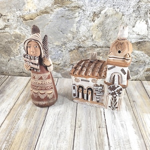 May include: A terracotta figurine of a person wearing a pointed hat and playing a pan flute, standing next to a terracotta building model featuring a tiled roof, a bell tower with a clock face, and three small figures visible in an arched doorway.