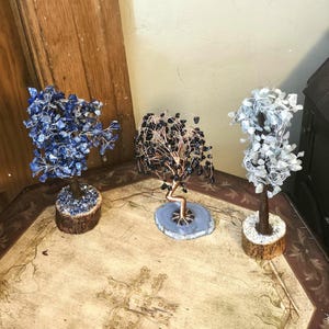 May include: Three decorative wire trees with gemstone leaves. The left tree has blue stones on a wood base, the center tree has black stones on a gray agate slice, and the right tree has clear white stones on a wood base.