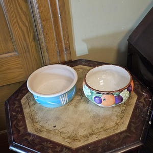 May include: Two ceramic bowls. The left bowl is light blue with white vertical stripes. The right bowl features a raised, colorful relief design of oranges and purple grapes around the exterior, with a glossy white interior finish.