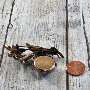 May include: A vintage silver-toned brooch with a floral design and a round watch face. The brooch is about the size of a US penny.