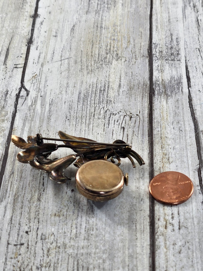 May include: A vintage silver-toned brooch with a floral design and a round watch face. The brooch is about the size of a US penny.