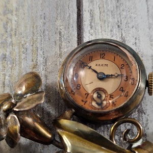 May include: A vintage gold-toned brooch featuring a floral design with a small pocket watch attached. The watch face is cream-colored with black hands and Roman numeral markings. The watch face also has the word "ELEM" printed on it.