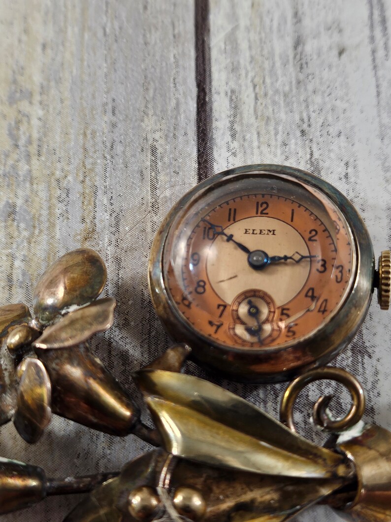 May include: A vintage gold-toned brooch featuring a floral design with a small pocket watch attached. The watch face is cream-colored with black hands and Roman numeral markings. The watch face also has the word "ELEM" printed on it.