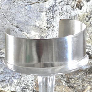 May include: A wide, polished silver-tone metal cuff bracelet with a smooth, reflective surface and a C-shaped open design. The bracelet features a clean, unadorned band that wraps around the wrist, showcasing a high-shine finish and a simple, structural form.
