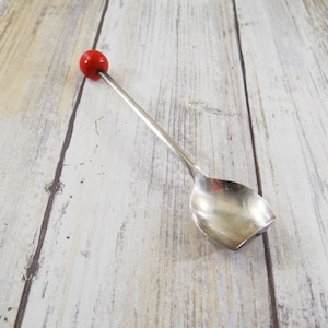 May include: A silver spoon with a rounded bowl and a long, slender handle. The handle is topped with a small, round, red bead. The spoon is likely used for serving or stirring. The spoon is on a light-colored wooden surface.
