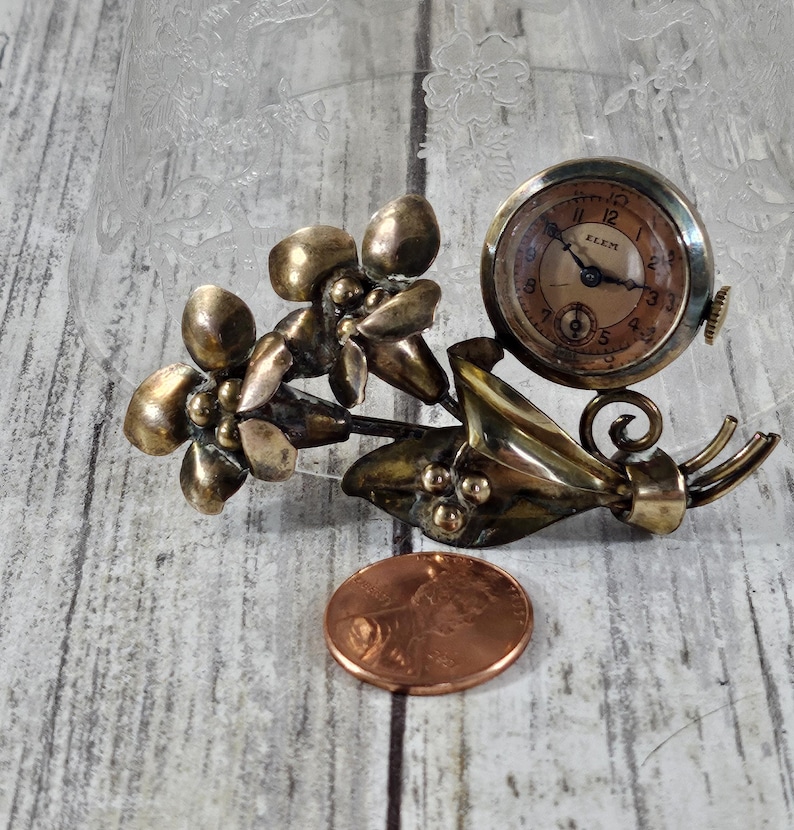 May include: A gold-toned brooch featuring a cluster of stylized flowers with a small, round watch attached. The watch face is cream-colored with black Roman numeral markings and the word "KLEM" printed on it.
