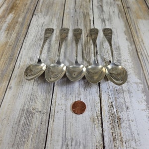 May include: Five silver spoons with ornate handle detailing are arranged on a light wood surface. A copper penny is placed near the spoons for scale. The spoons have a reflective surface and a classic design.