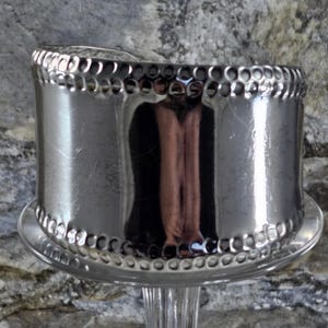 May include: A silver napkin ring with a hammered design around the top and bottom edges. The napkin ring is cylindrical and sits on a clear glass stand.