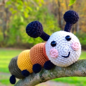 Puede incluir: Un juguete de peluche de oruga hecho a crochet con una cabeza blanca, ojos negros y mejillas rosadas. El cuerpo está segmentado con secciones marrones, amarillas y verdes. La oruga está sentada en una rama.