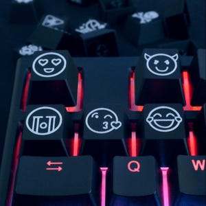May include: A black mechanical keyboard with custom emoji keycaps. The keycaps display white emoji designs such as hearts and a smiling devil face. The keyboard has red backlighting, creating a striking visual effect.