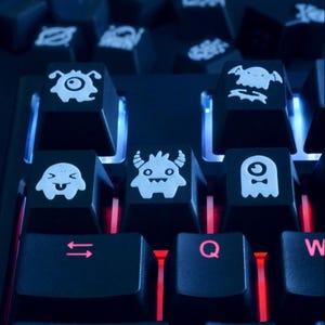 May include: A close-up of a black keyboard with custom keycaps. The keycaps have white cartoon monster designs. The keys are lit with blue and red lights. A fun and unique accessory for a computer.