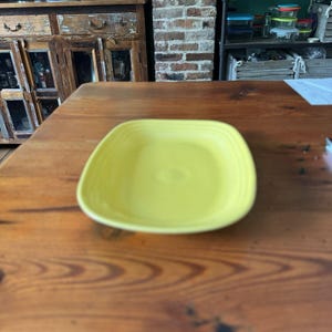 May include: A square, yellow ceramic plate with rounded corners. The plate is centered on a wooden table. The plate has a slightly raised rim and a smooth, glossy surface. The plate is a bright, cheerful yellow color.