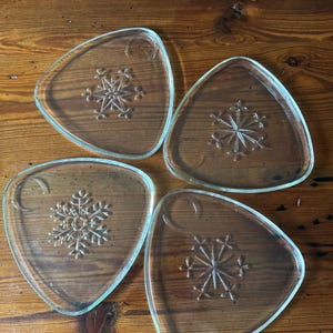 May include: Four clear glass plates with a triangular shape and a snowflake design in the center. Each plate has a curved design in the upper corner. The plates are on a wooden surface.