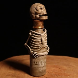 Puede incluir: Una botella decorativa con un diseño de esqueleto. La botella tiene un tapón en forma de calavera y un torso de esqueleto envuelto alrededor de la botella. El texto "SKELE-V" es visible en la botella. La botella se exhibe sobre una superficie de madera.