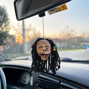 以下が含まれることがあります： 茶色の顔と黒いドレッドロックが付いた車の芳香剤。顔には笑顔が描かれており、ドレッドロックには緑と黄色のビーズが付いています。