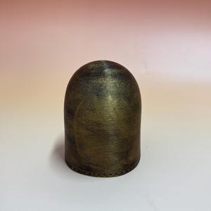 May include: A cylindrical, domed object with a metallic gold and black finish. The object has a textured surface and a series of small holes near the base. The background is a gradient of pink and white.