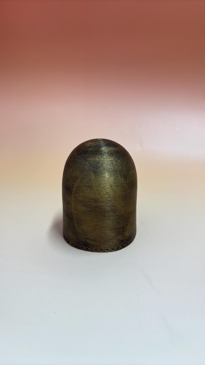 May include: A cylindrical, domed object with a metallic gold and black finish. The object has a textured surface and a series of small holes near the base. The background is a gradient of pink and white.